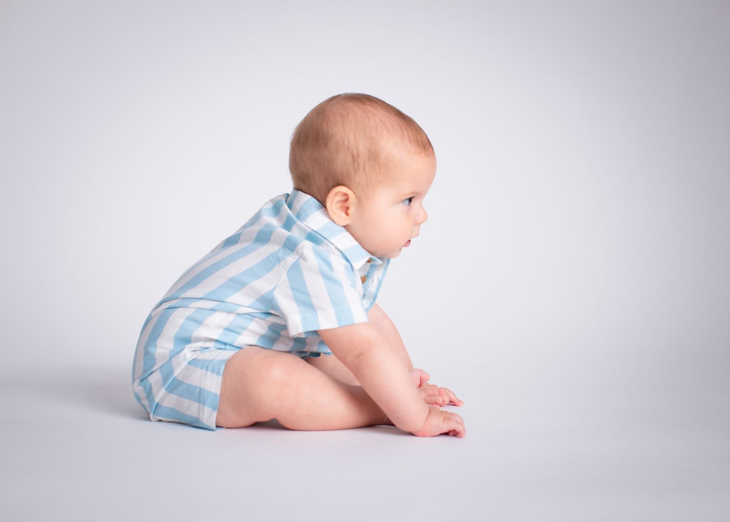Baby in blue striped onesie sitting and looking to the side on white background.