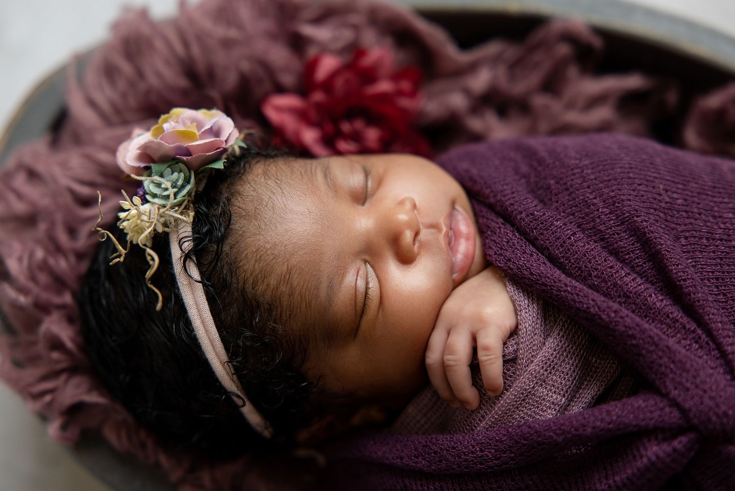 Peaceful newborn baby swaddled in purple wrap with floral headband, sleeping in a bowl with soft purple fabric.