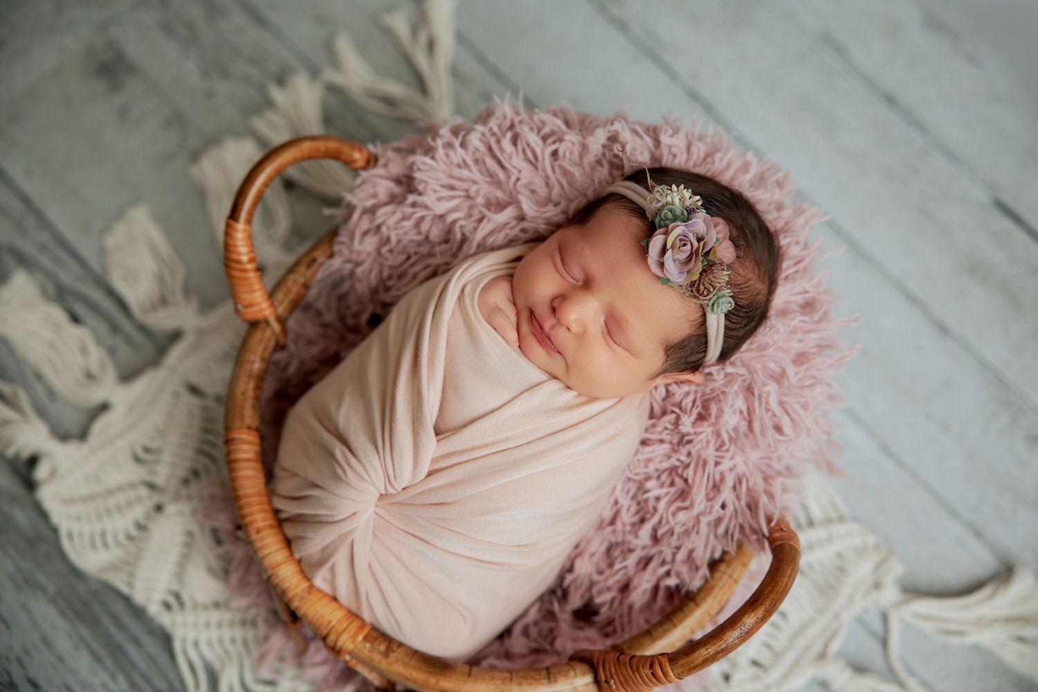 Newborn baby swaddled in pink wrap sleeping in wicker basket with floral headband and fluffy pink fur.