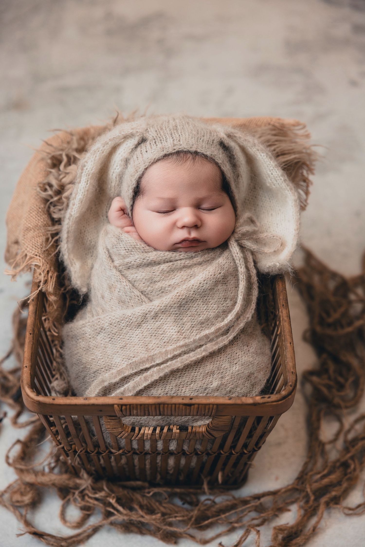 Sleeping newborn baby swaddled in beige wrap wearing bunny ear hat, resting in rustic wicker basket.