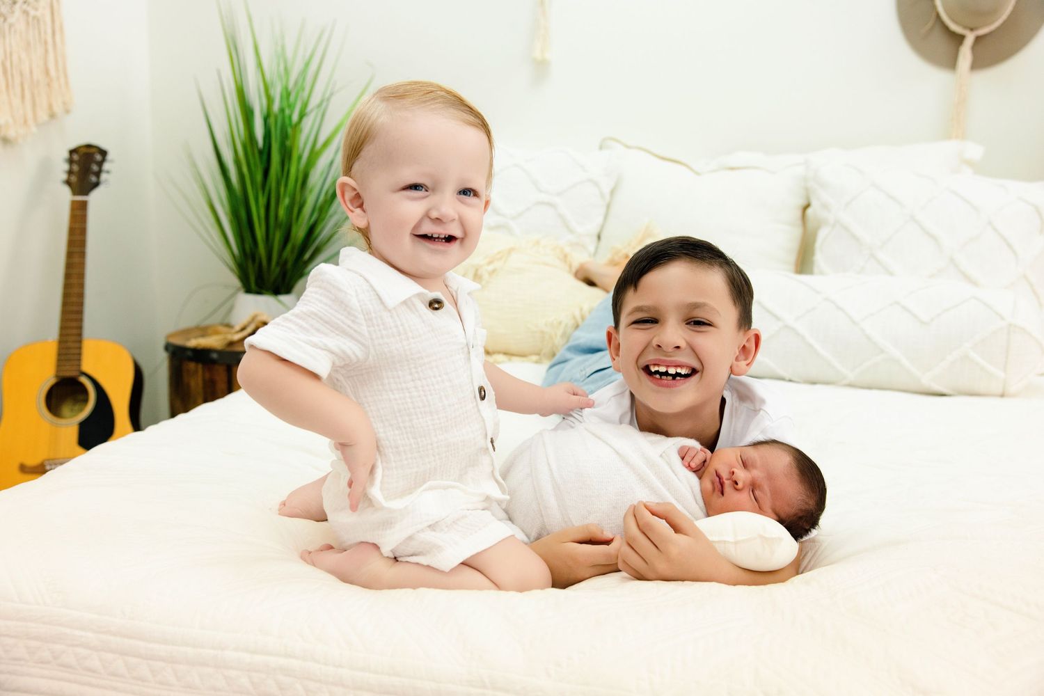 Two smiling boys pose on a white bed with a newborn baby, with a guitar and plant in the background.
