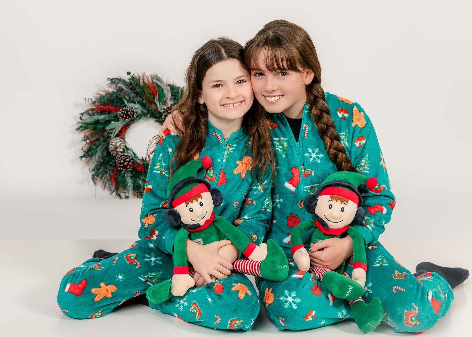 Two girls in matching green Christmas pajamas holding elf dolls, sitting together with a holiday wreath backdrop.
