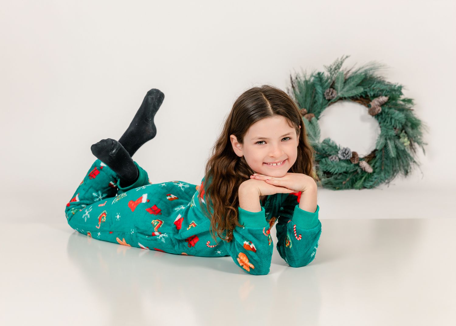 Girl in green Christmas pajamas lying down smiling, with a festive wreath in the background.