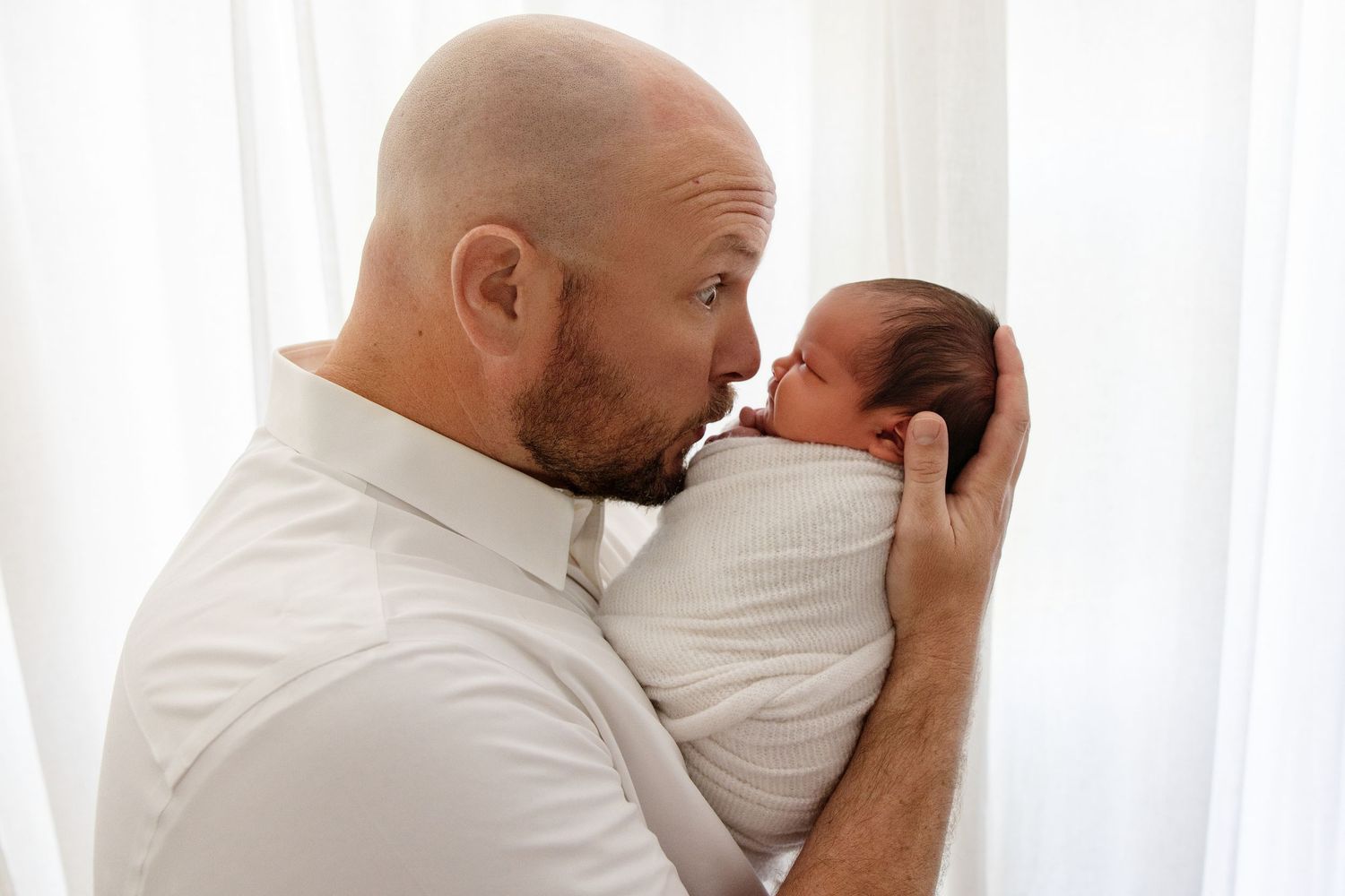 Bald father tenderly kissing his swaddled newborn baby, sharing a precious bonding moment near a bright window.