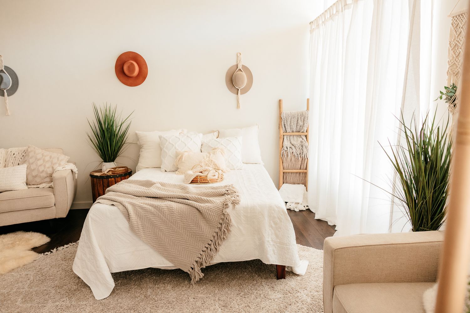 Bright boho bedroom with white bedding, decorative hats, ladder shelf, sheer curtains, and tall plants. in the Bonsall Studio near Fallbrook
