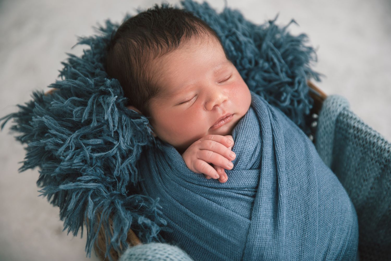 Peaceful newborn baby boy wrapped in teal blue blanket sleeping in a rustic wooden bowl.