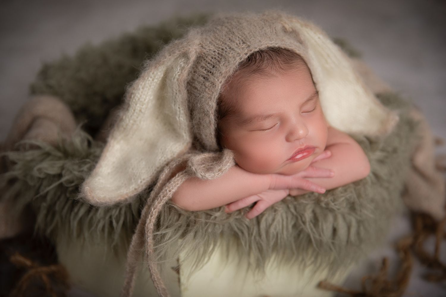 Sleeping newborn baby wearing knit bunny ear bonnet posed in green fur-lined bucket with chin on hands at The Studio Bonsall, near Fallbrook