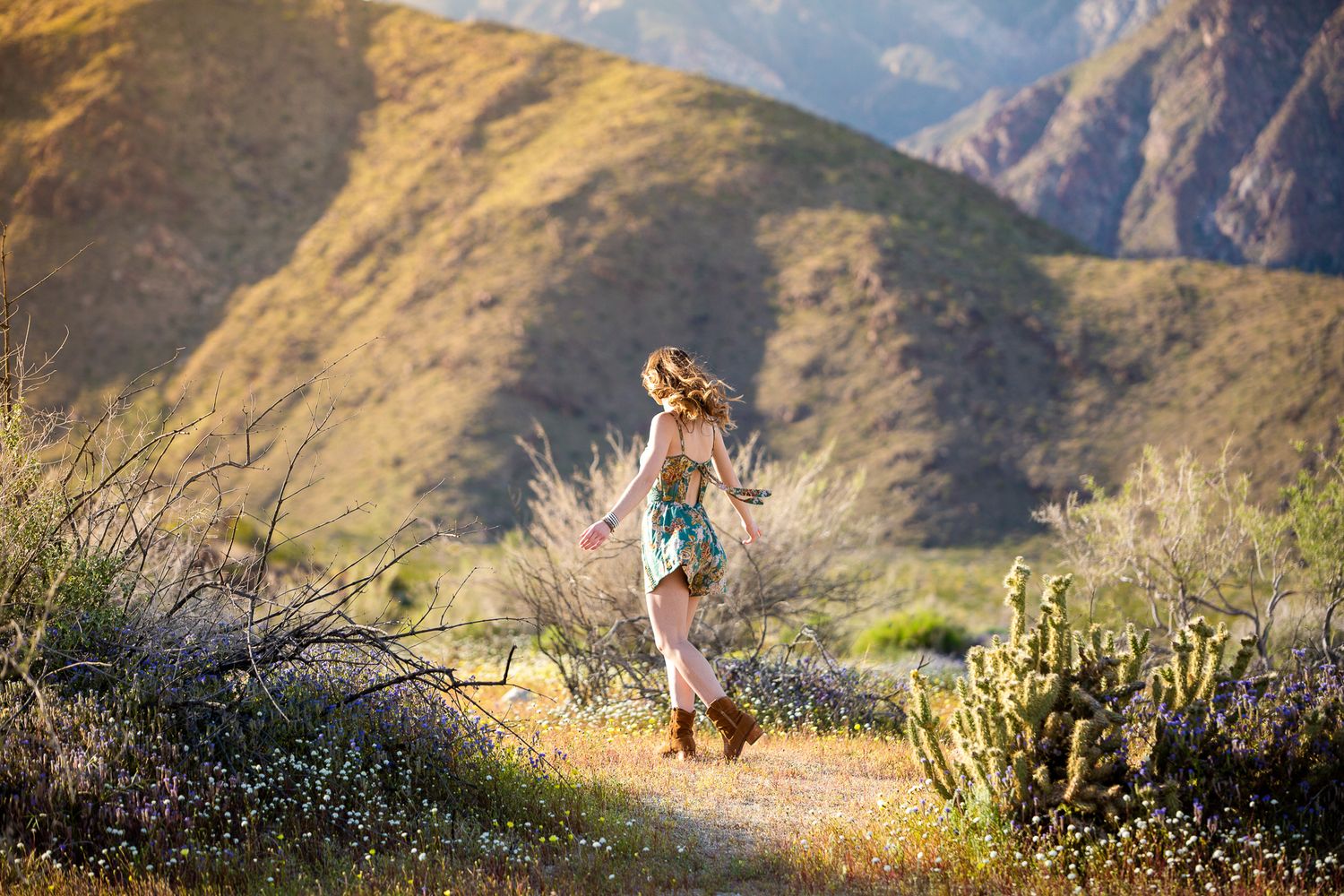 Young woman in floral dress walks along desert trail surrounded by mountains and cacti. Senior photography session in Vista CA for hig