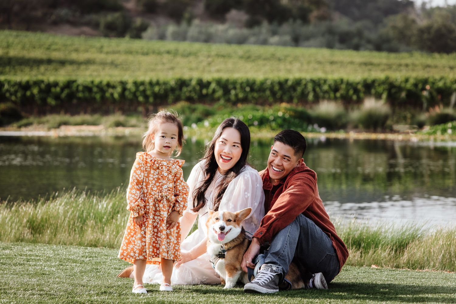 Smiling Asian family sits on grass with their corgi dog near a scenic vineyard pond.