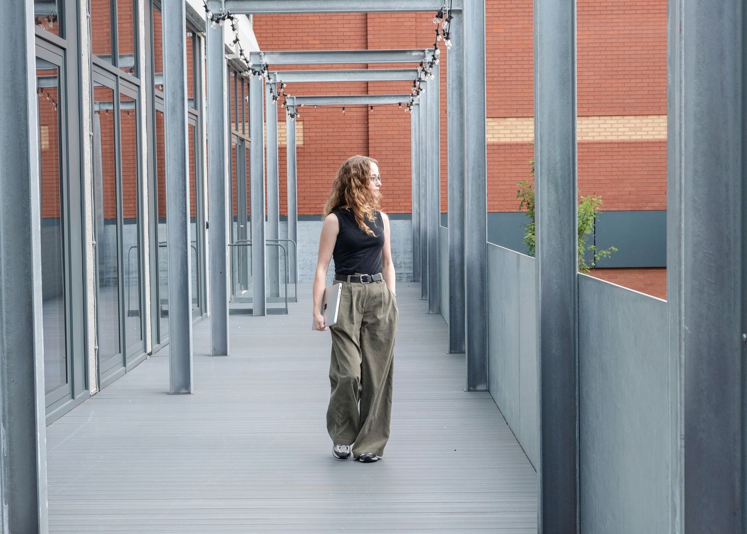 Woman in wide-leg khaki pants and black top walking through a modern covered walkway with gray metal columns.