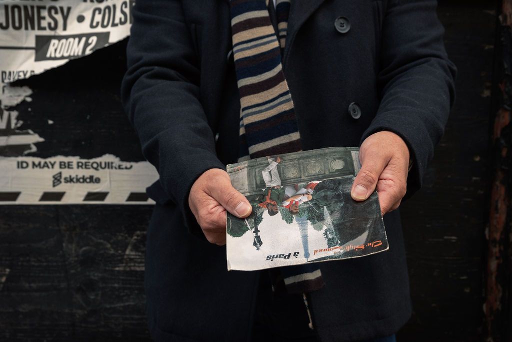 Man in a black coat holding a vintage photograph outdoors near a venue sign.