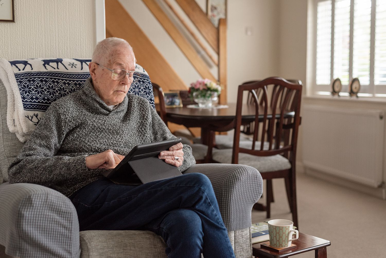 Elderly man sitting in armchair at home using a digital tablet in a cozy living room.