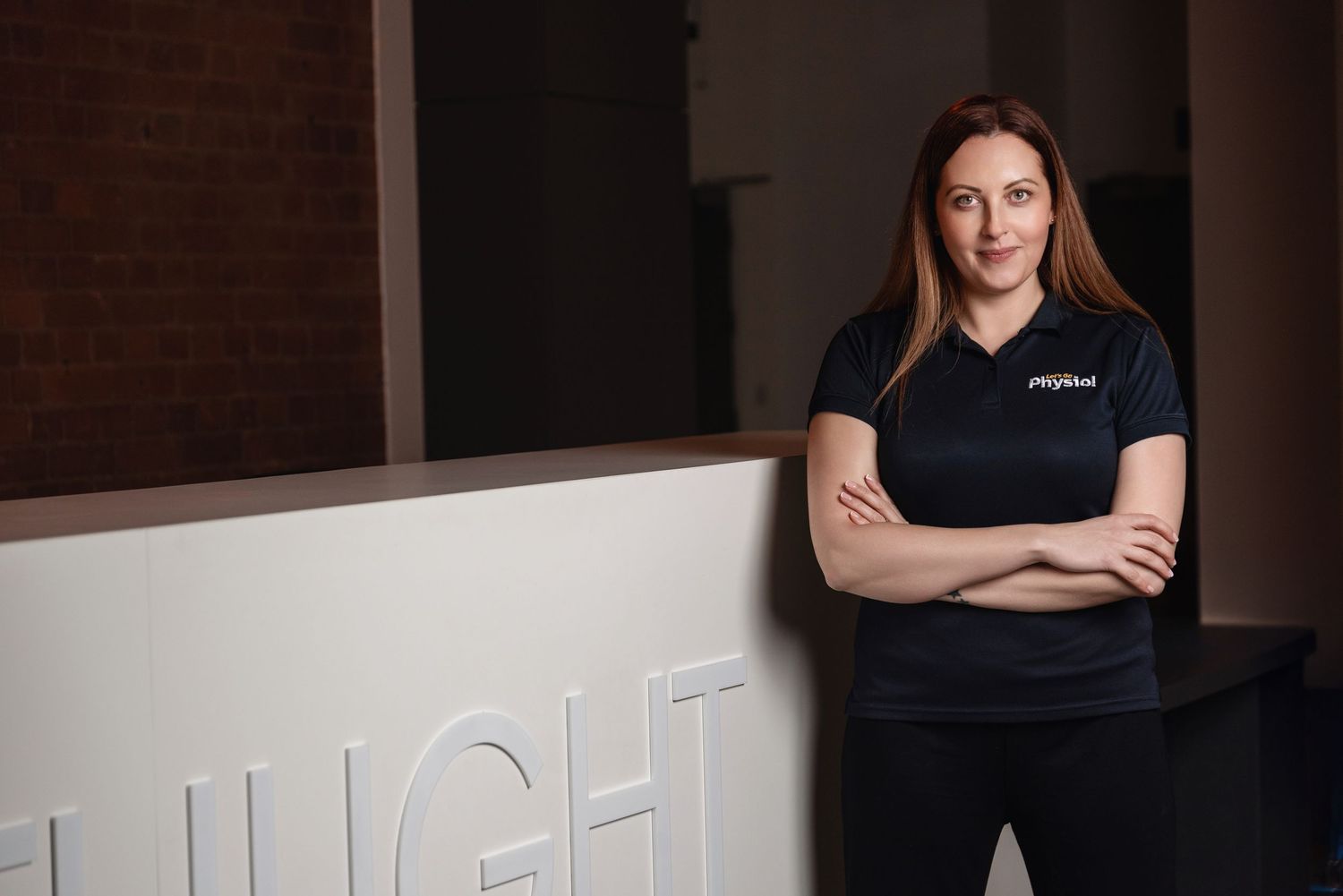 A confident woman in a black polo shirt stands with arms crossed in a modern lobby with illuminated signage.