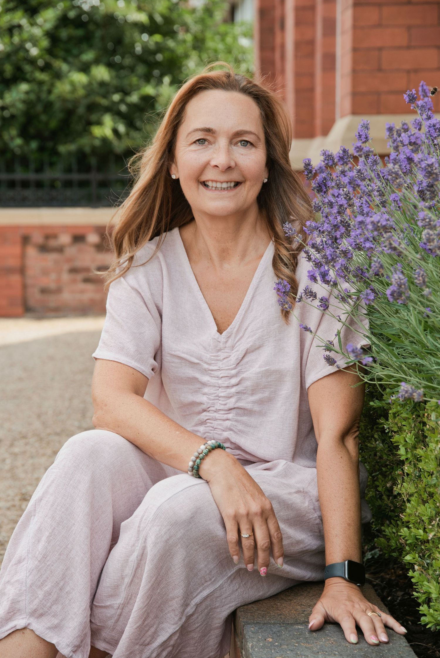 Smiling middle-aged woman in pink linen outfit sitting outdoors near purple lavender flowers by a brick building.