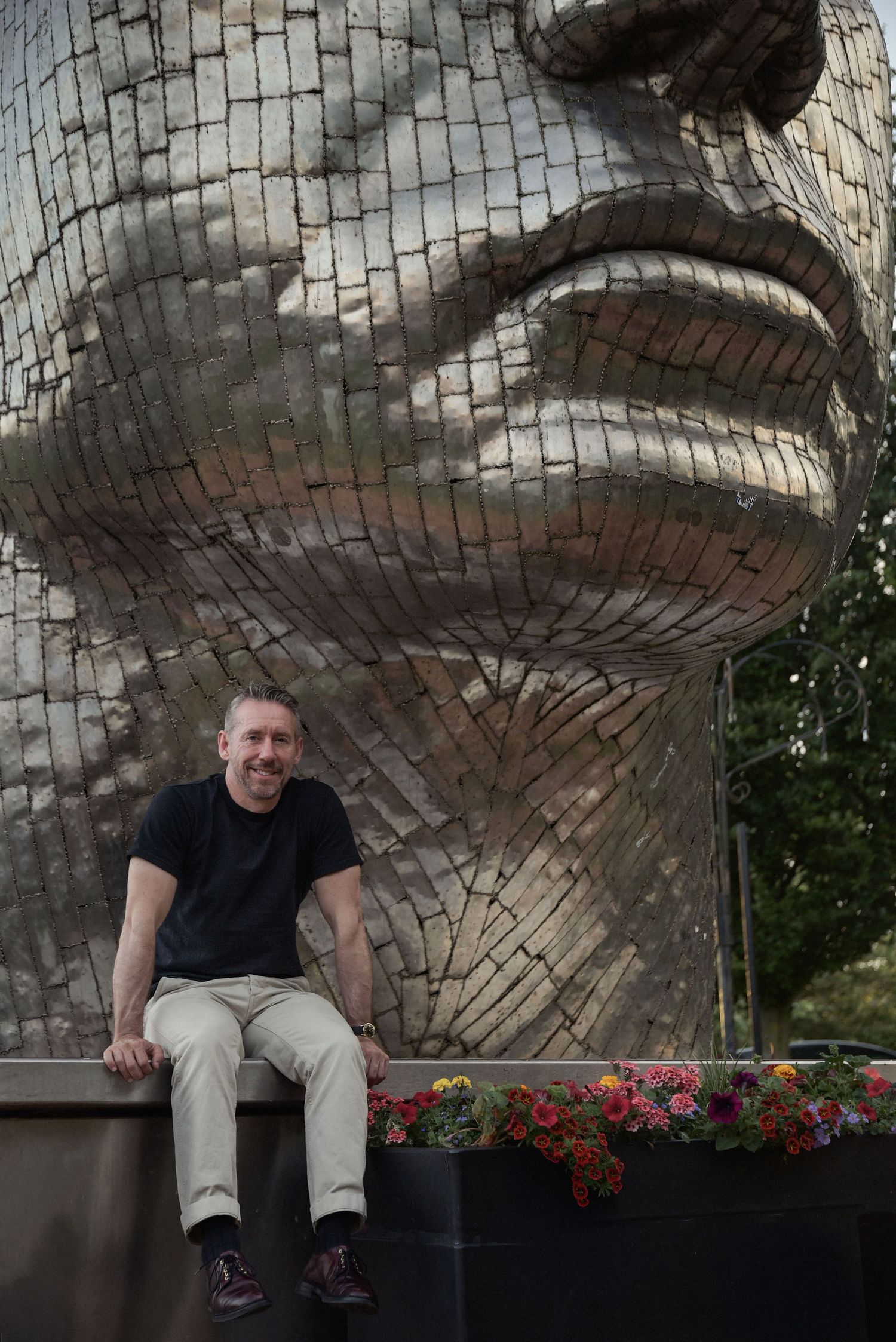 Man smiling in front of a large mosaic face sculpture outdoors, surrounded by colorful flowers.