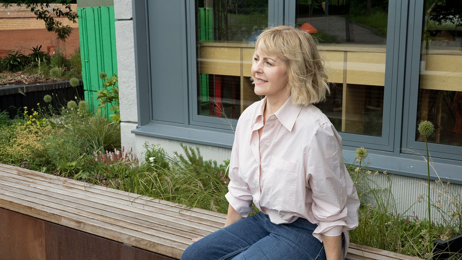 Woman with blonde hair in pink shirt and jeans sits smiling on wooden bench beside green planters outdoors.
