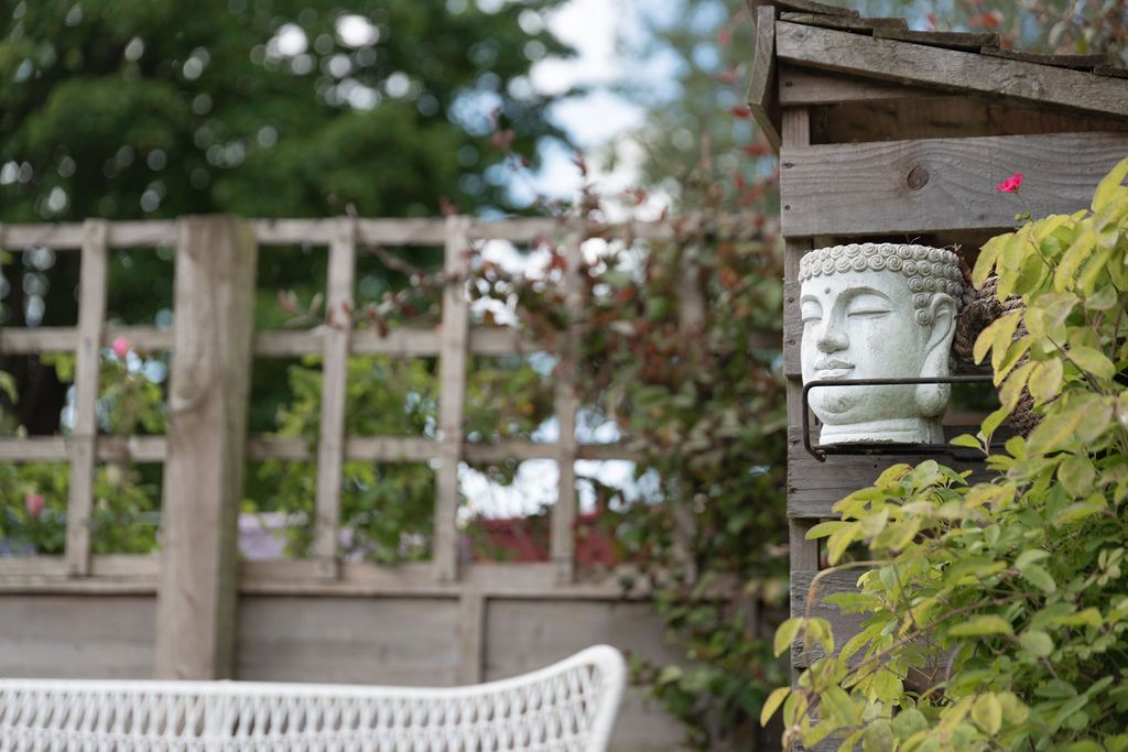 Buddha head wall planter mounted on wooden fence in lush garden with white wicker chair and green foliage.