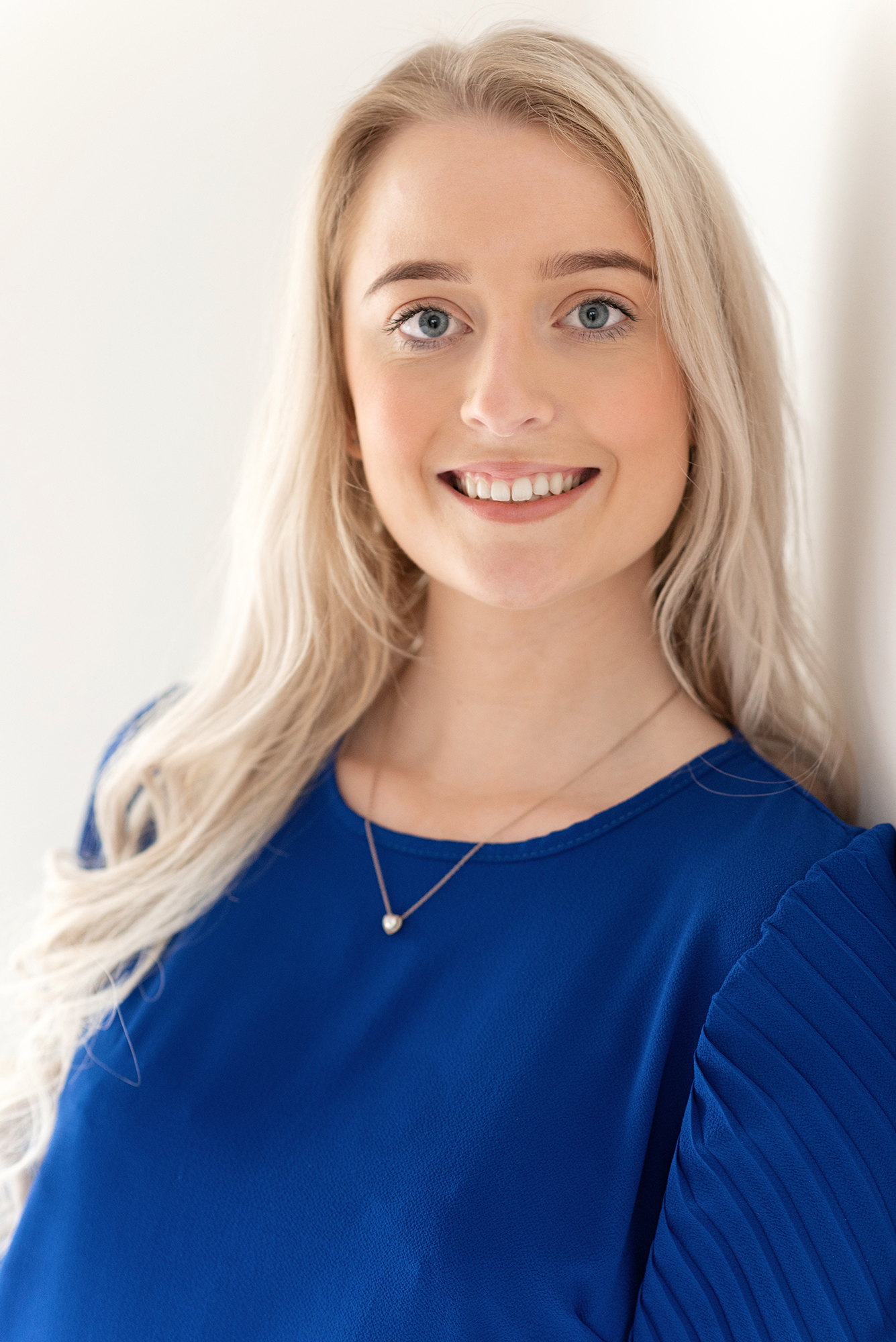 Professional portrait of woman wearing royal blue top and delicate necklace against white background.