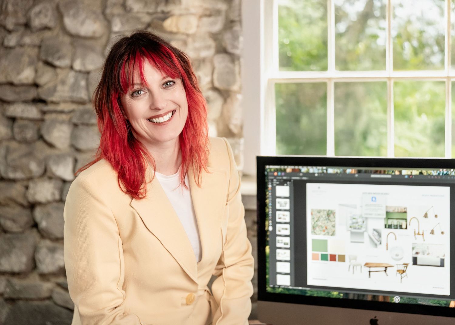 Interior designer with bright red hair smiles while presenting a design layout on computer monitor near stone wall.