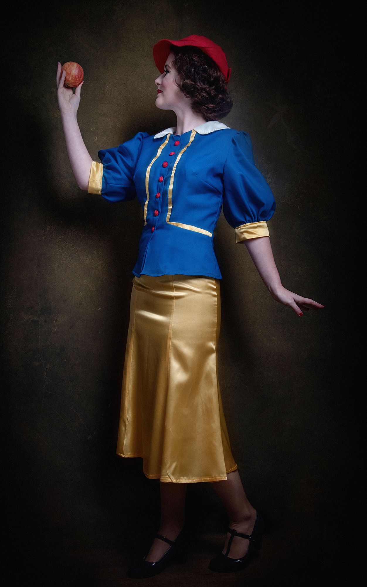 A vintage-inspired costume with blue top, gold skirt, red hat, and apple prop.