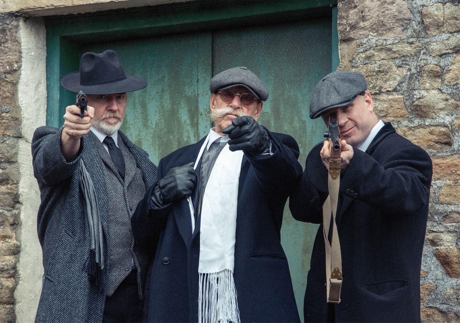 Three figures in vintage 1920s attire with hats stand against a stone wall with green door.