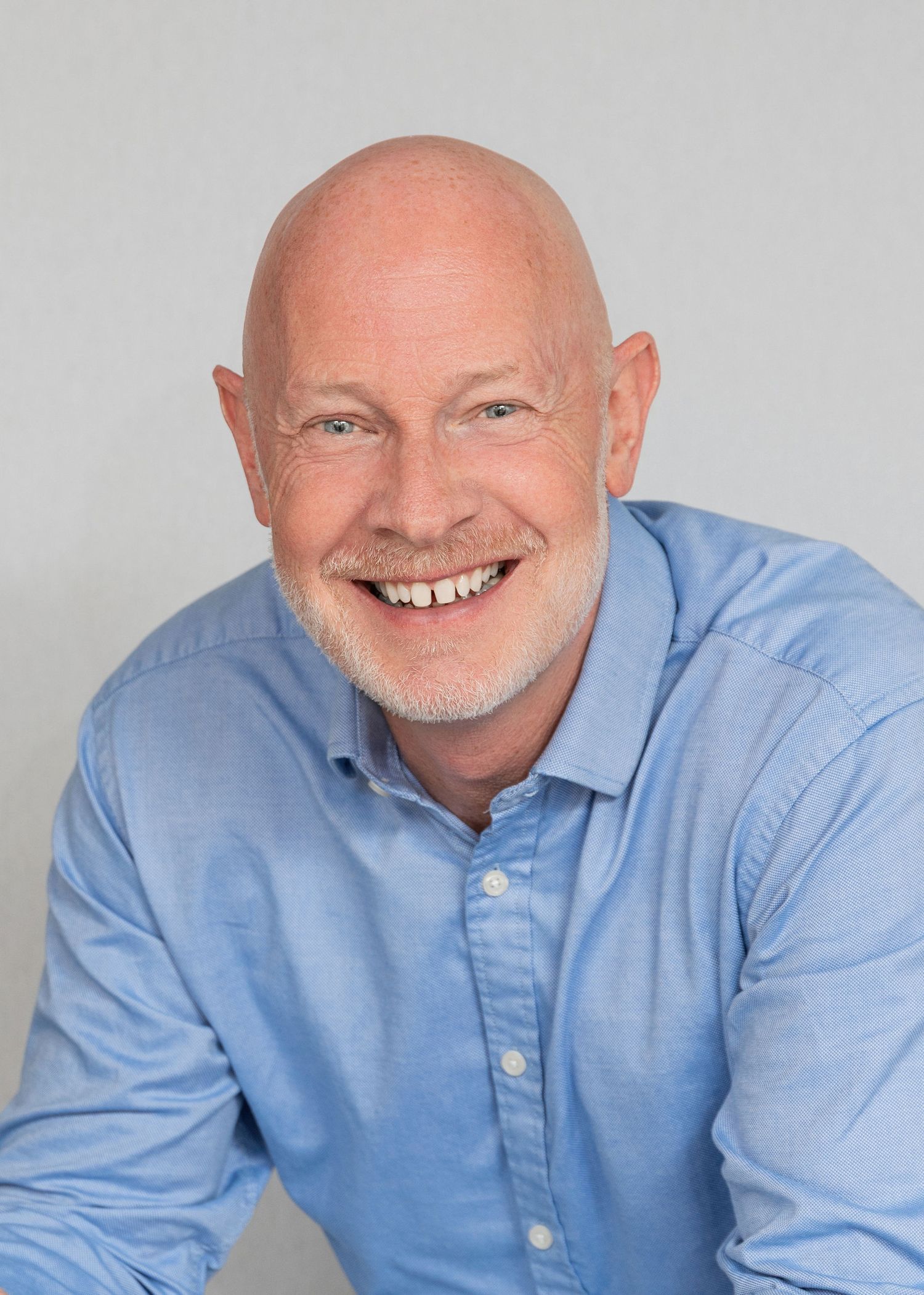 Smiling bald man with gray beard wearing a light blue shirt against a gray background.