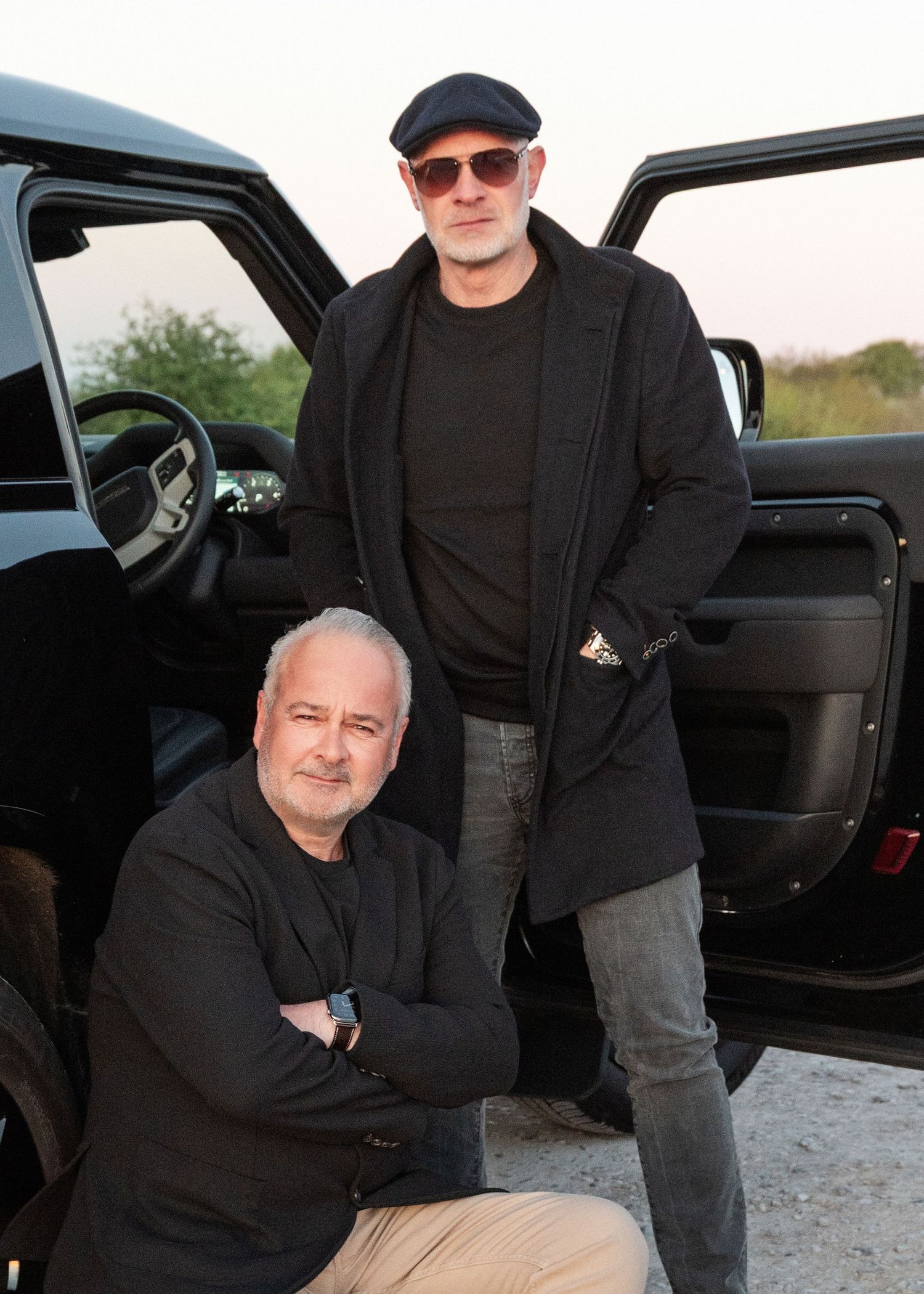 Two men in dark clothing posing beside a black SUV outdoors at dusk.