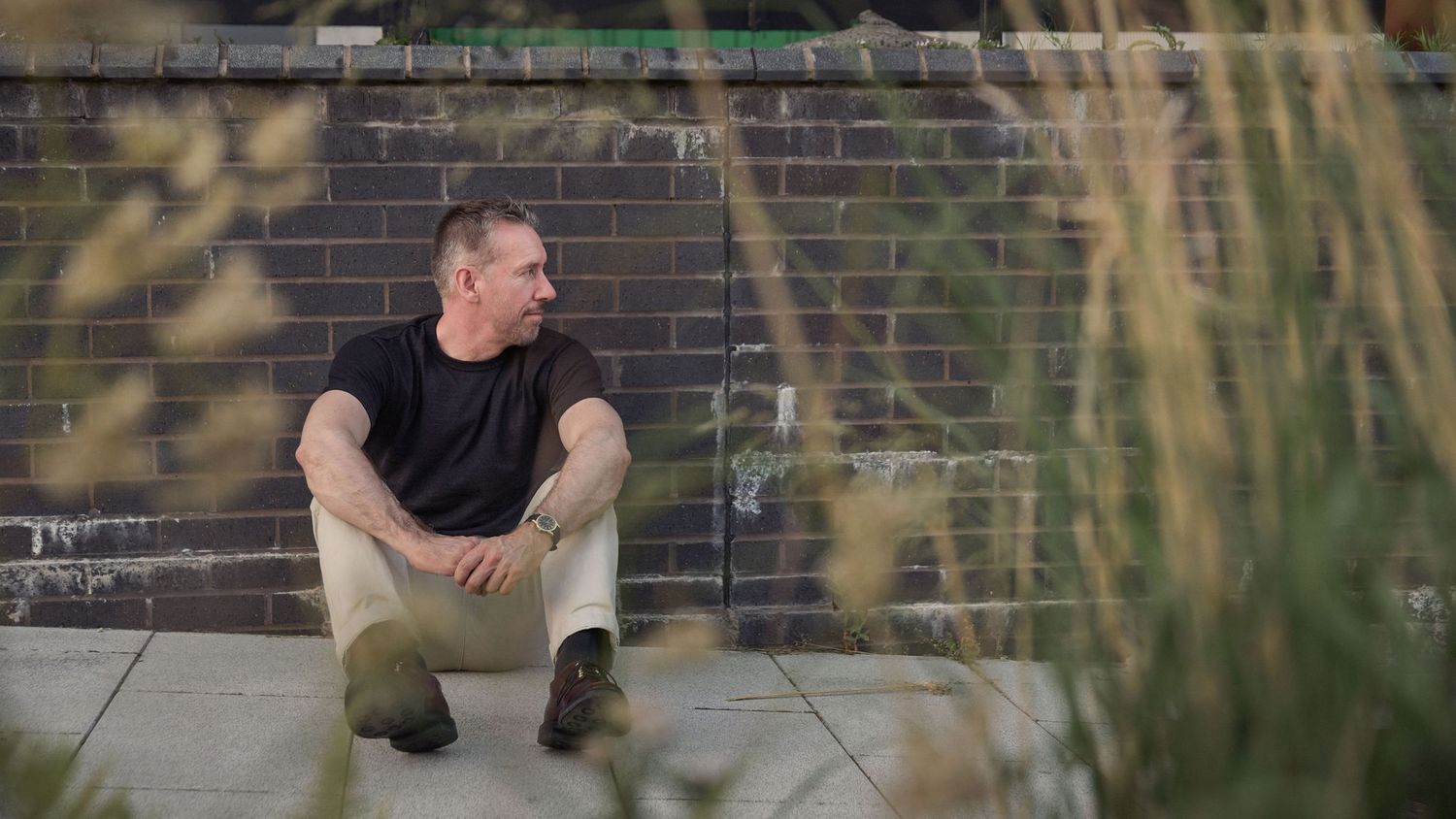 Man in black t-shirt sitting against a graffiti brick wall outdoors, surrounded by tall grass.