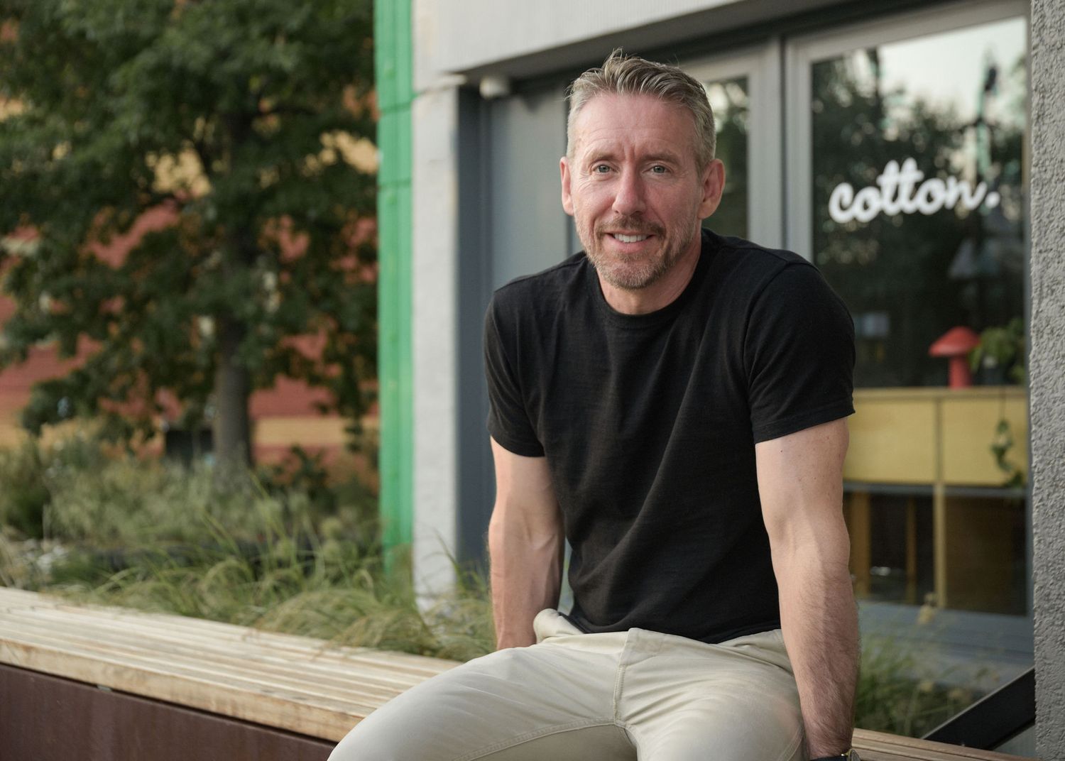 A smiling middle-aged man with grey hair sits outdoors in a black t-shirt and khaki pants near a modern building.
