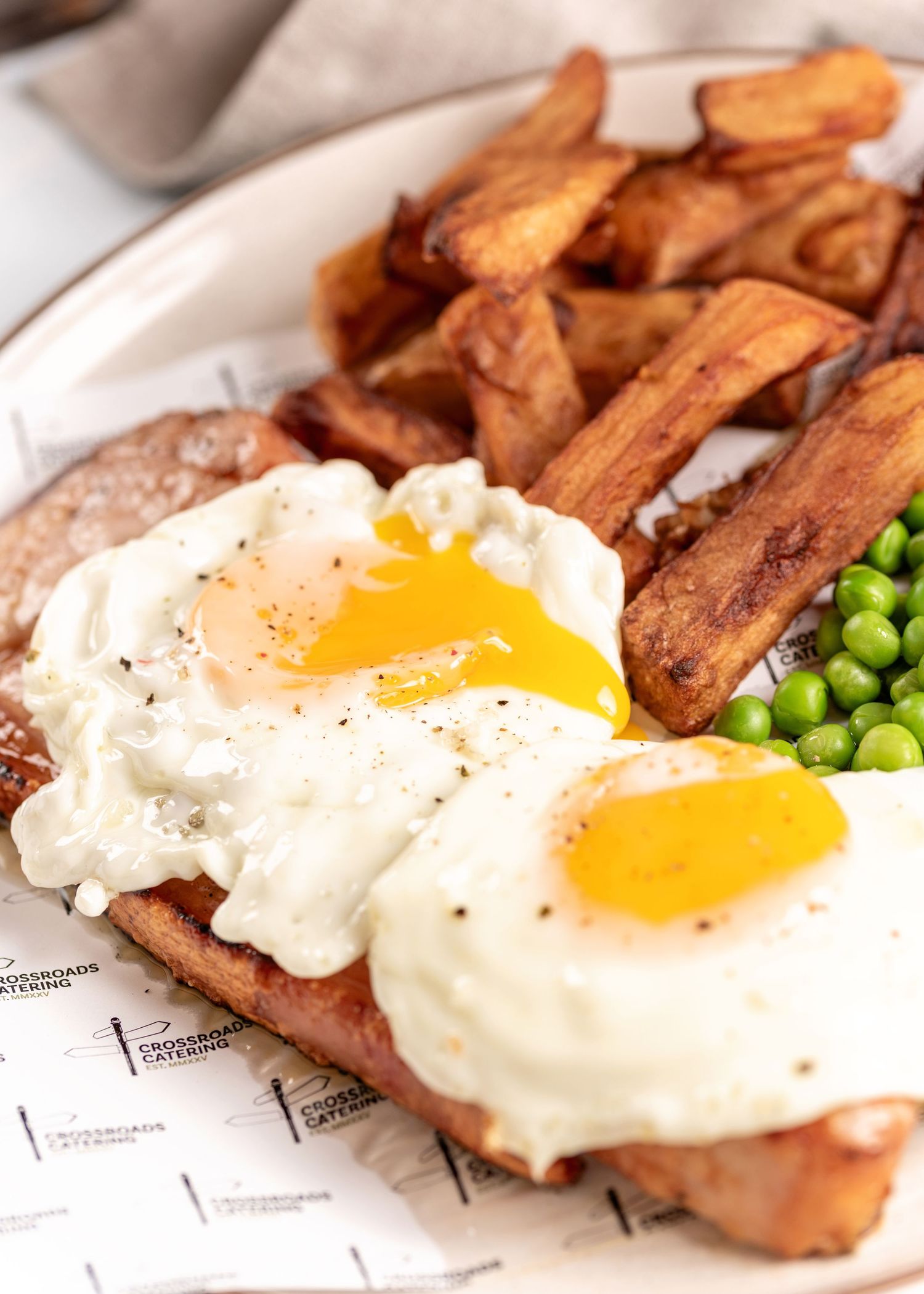 Plate of two fried eggs on meat with thick-cut chips and green peas, served on Crossroads Catering branded paper.