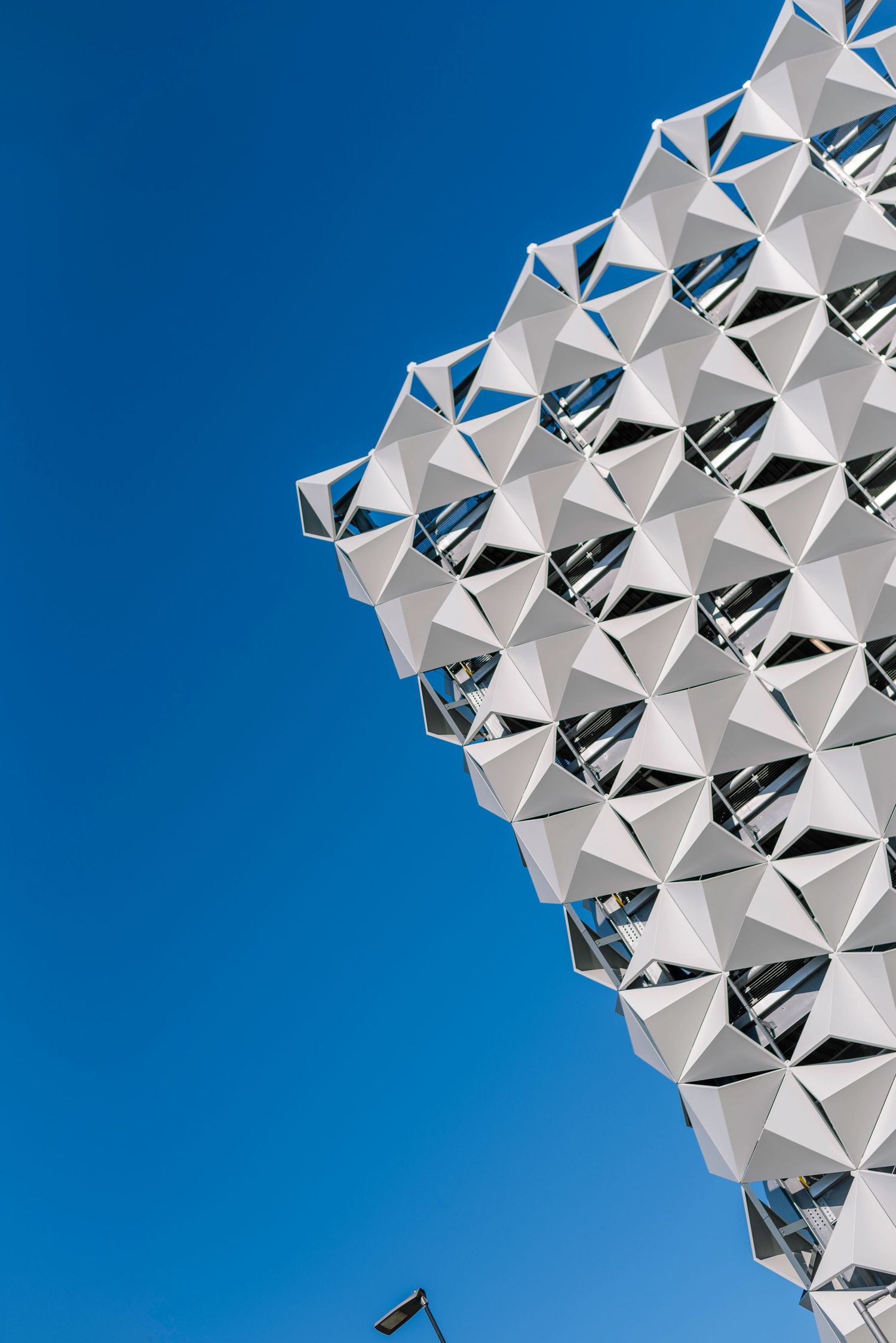Modern building facade with geometric white triangular panels against a clear blue sky, shot from below.