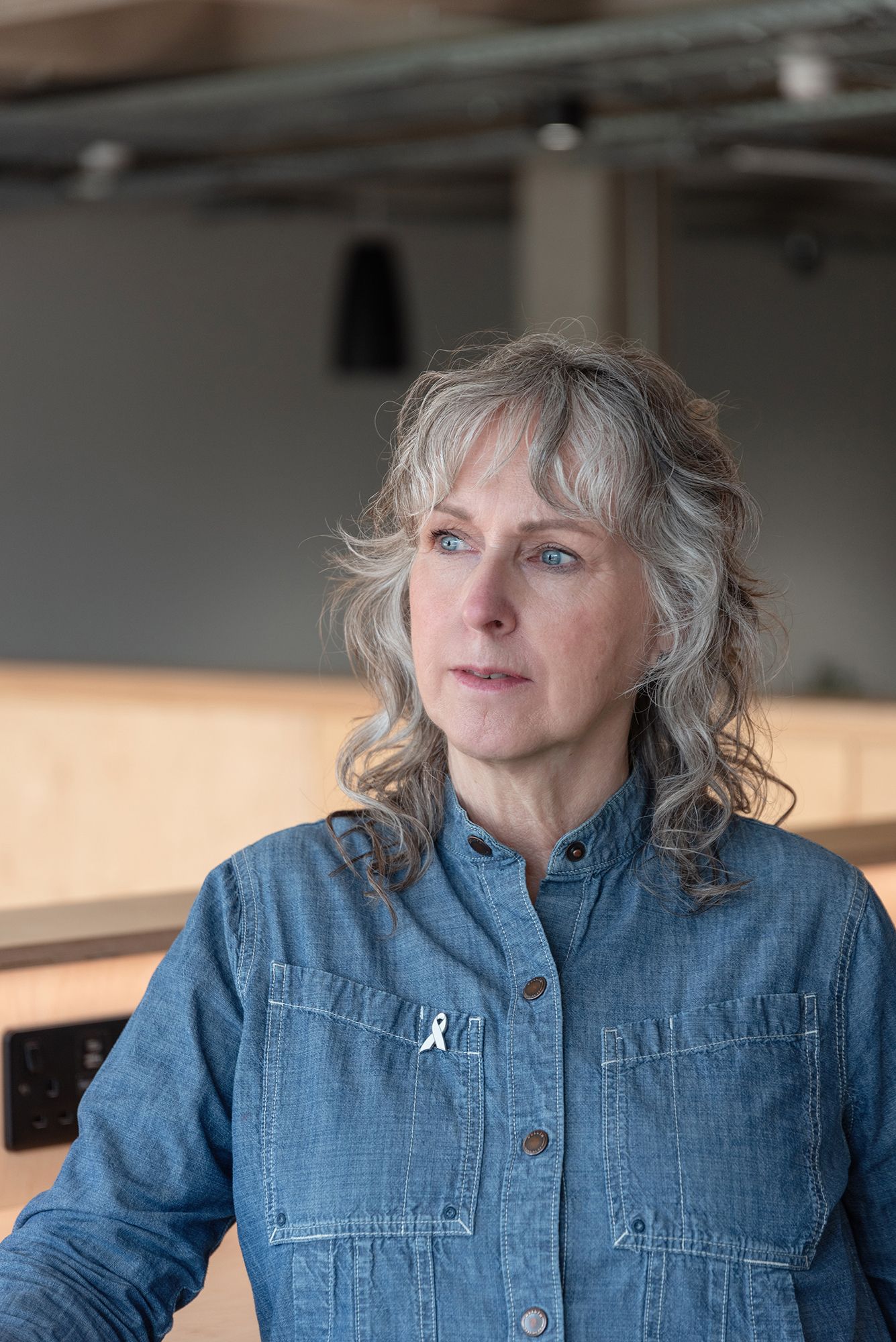 Middle-aged woman with gray hair and blue eyes wearing a denim shirt with a white ribbon pin indoors.