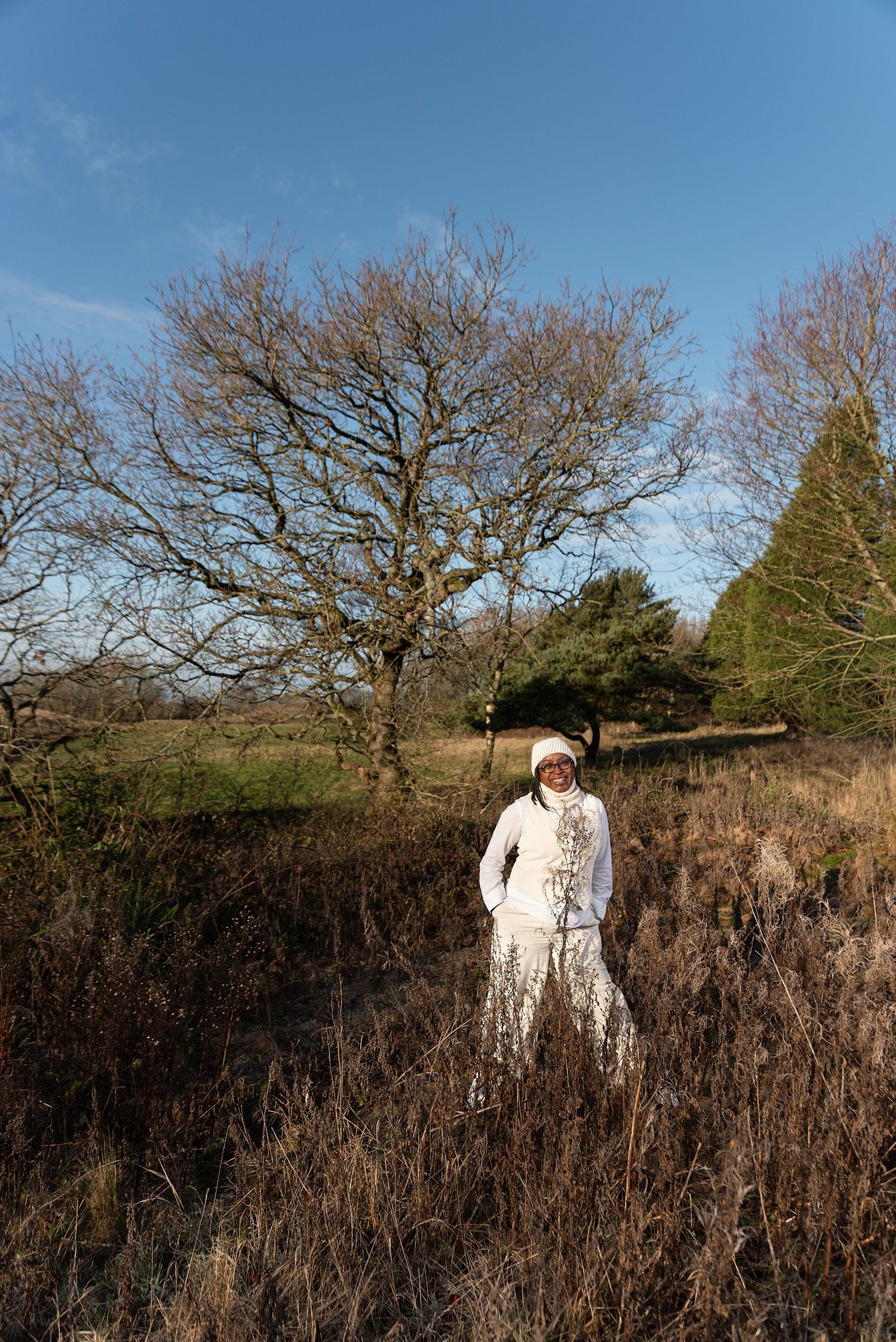 A person in a white winter coat stands among tall brown dried grass and bare winter trees in a rural setting.