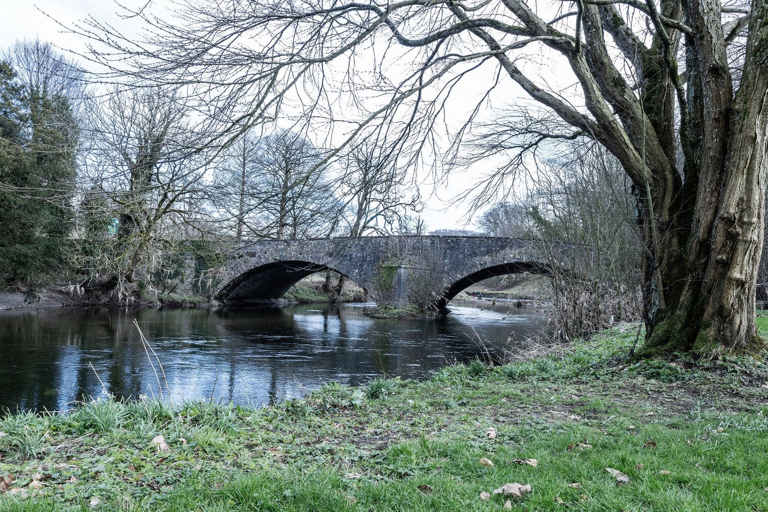 Historic stone bridge spanning over a river with bare winter trees in the foreground.