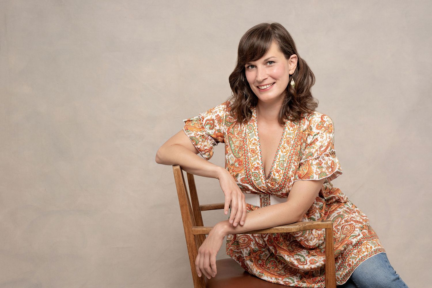 Portrait of person in floral dress sitting casually on wooden chair against neutral background.