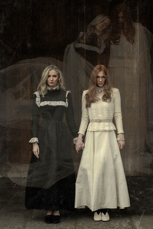 Two women in Victorian-style dresses, one in black and one in white, stand before a ghostly backdrop.