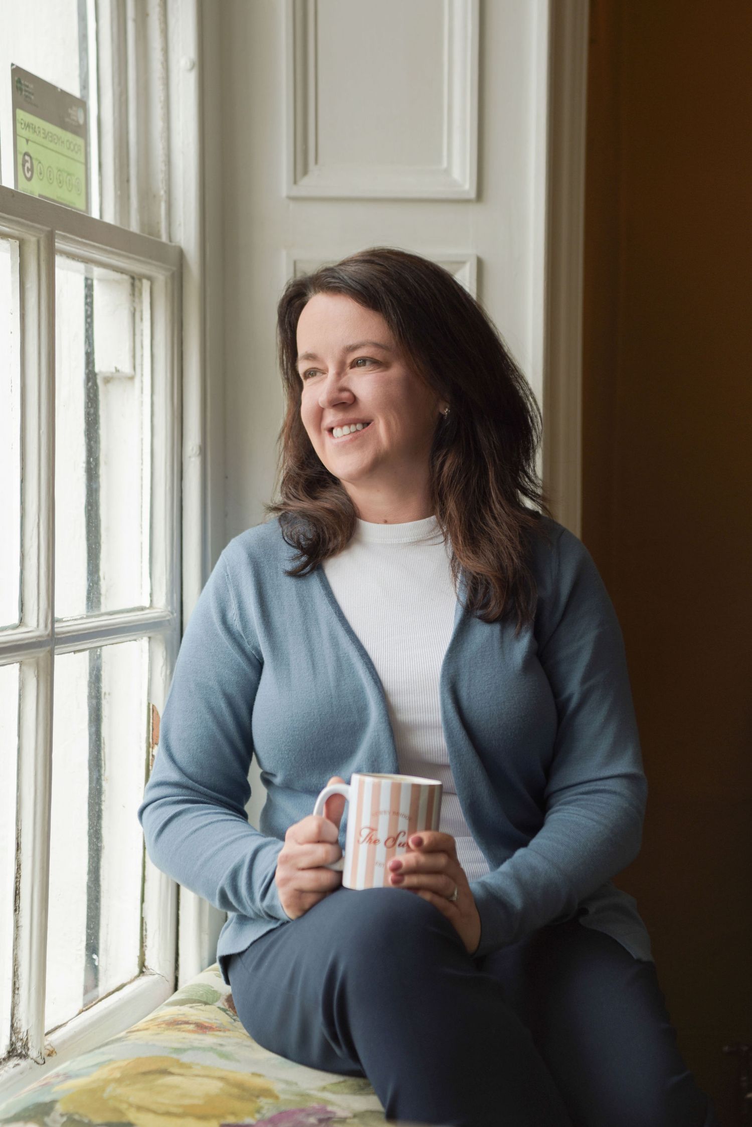 Person in blue cardigan sits by window holding white mug while smiling and looking to the side.