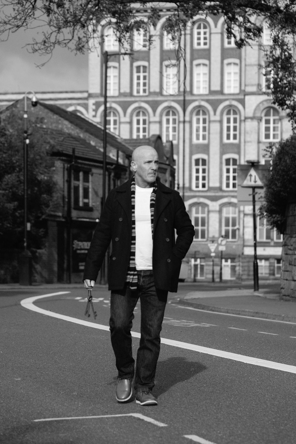 Bald man in jacket and jeans stands on an empty road with a historic building in black and white.