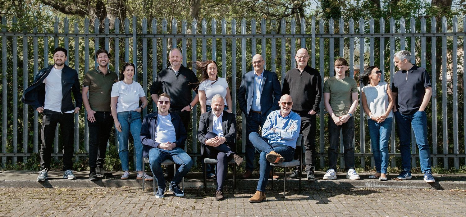 A diverse group of professionals posing outdoors in front of a metal fence on a sunny day.