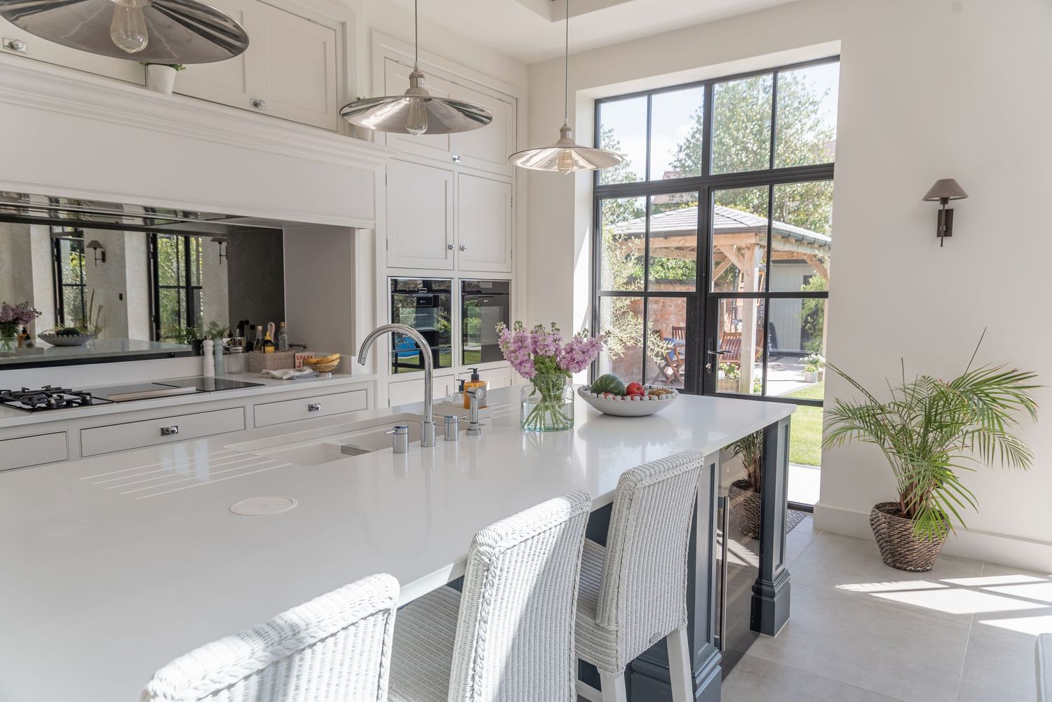 Bright modern white kitchen with large island, pendant lights, and floor-to-ceiling steel-framed windows overlooking a garden.
