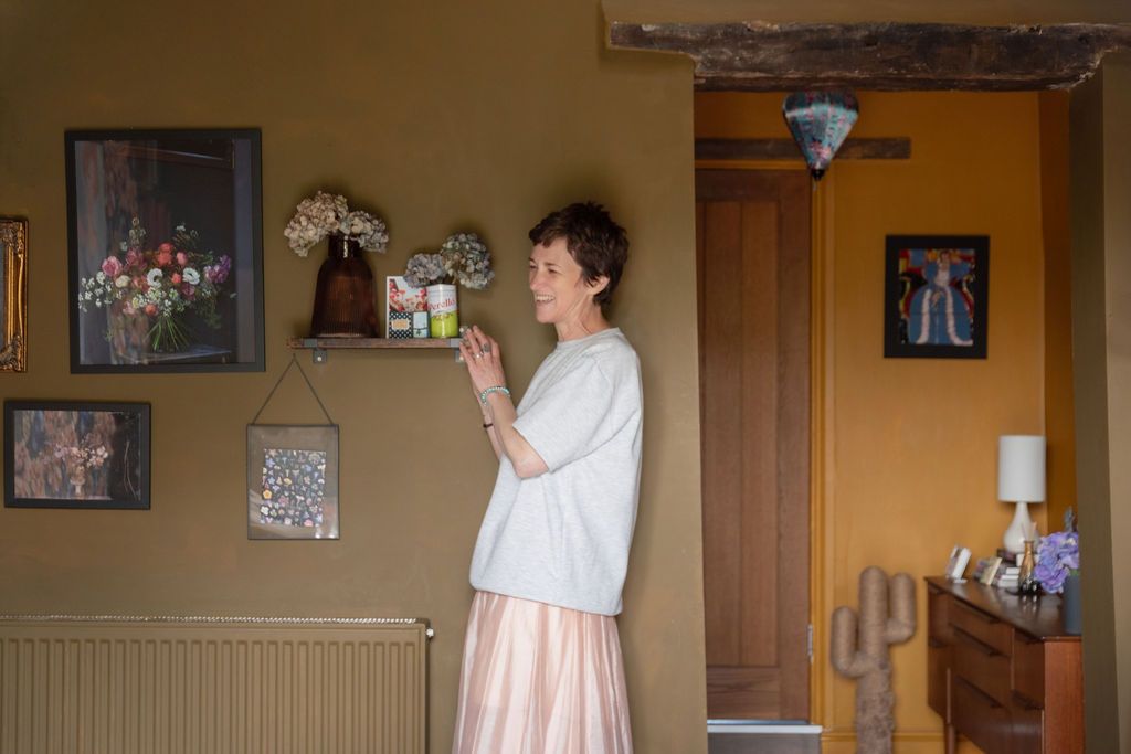Woman in white top and pink skirt stands in a warmly decorated room with framed artwork and a wooden shelf.