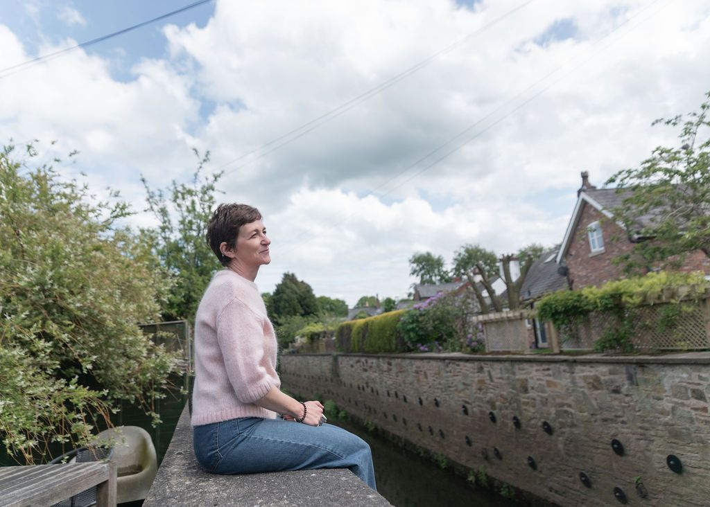Woman in pink sweater sitting on stone wall by a canal with lush greenery and historic buildings behind her.