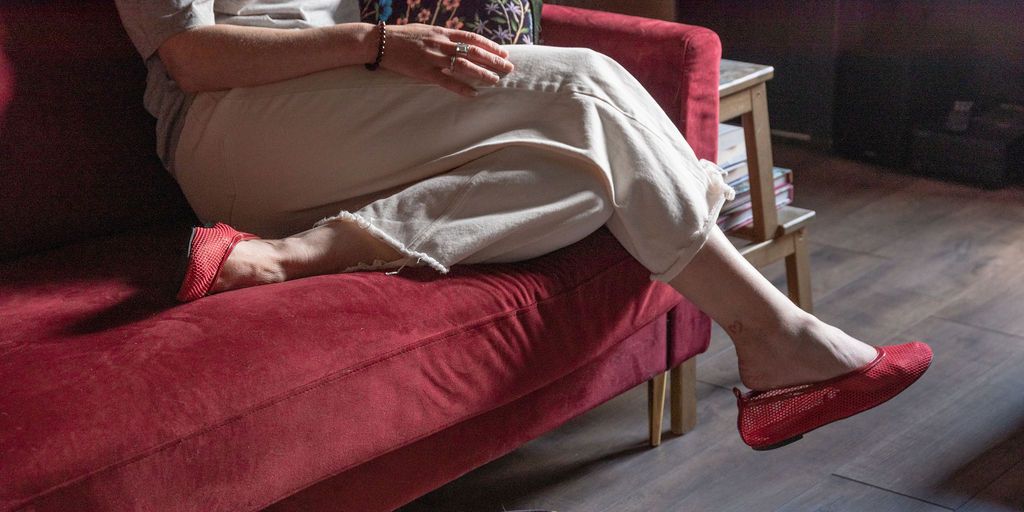 Woman relaxing on red velvet sofa wearing white pants and red flat shoes, legs crossed in cozy living room.