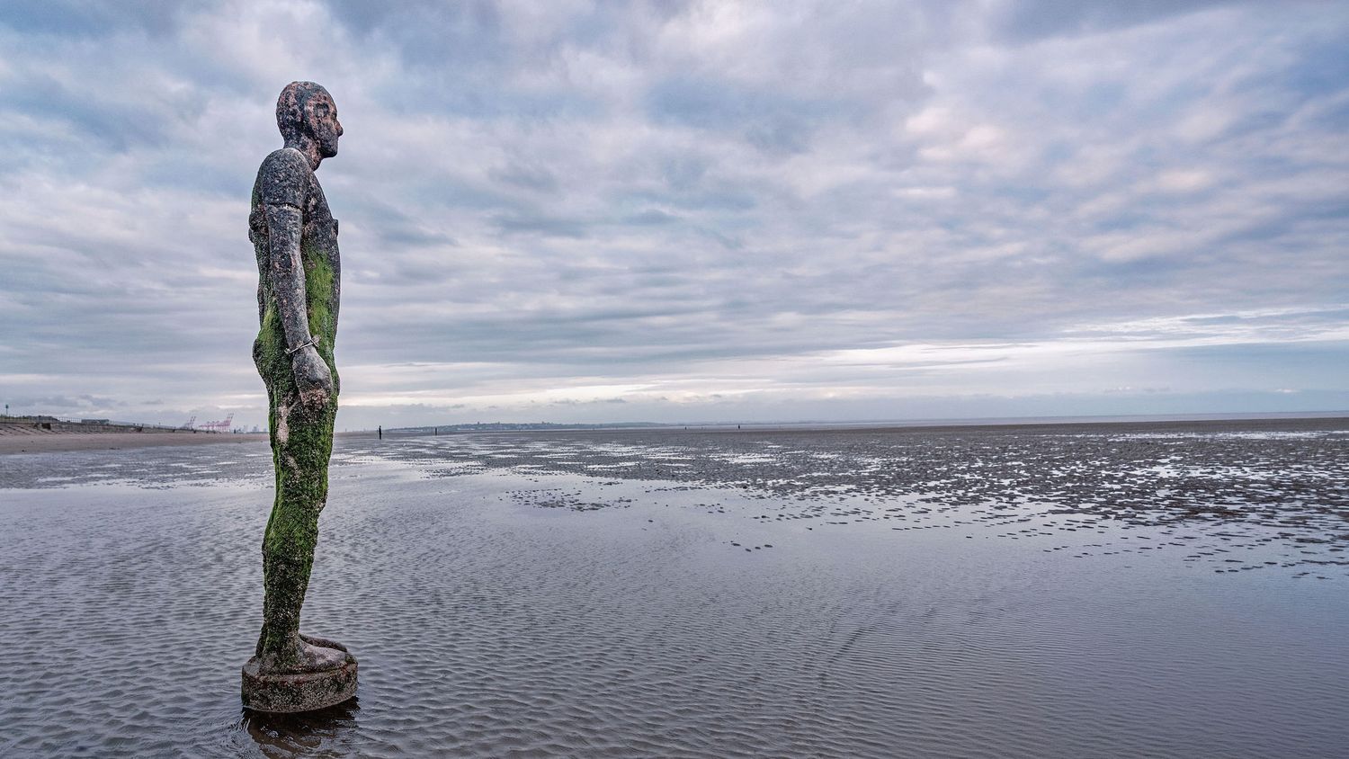 Iron man sculpture standing on Crosby Beach at low tide with dramatic cloudy sky reflected in wet sand.