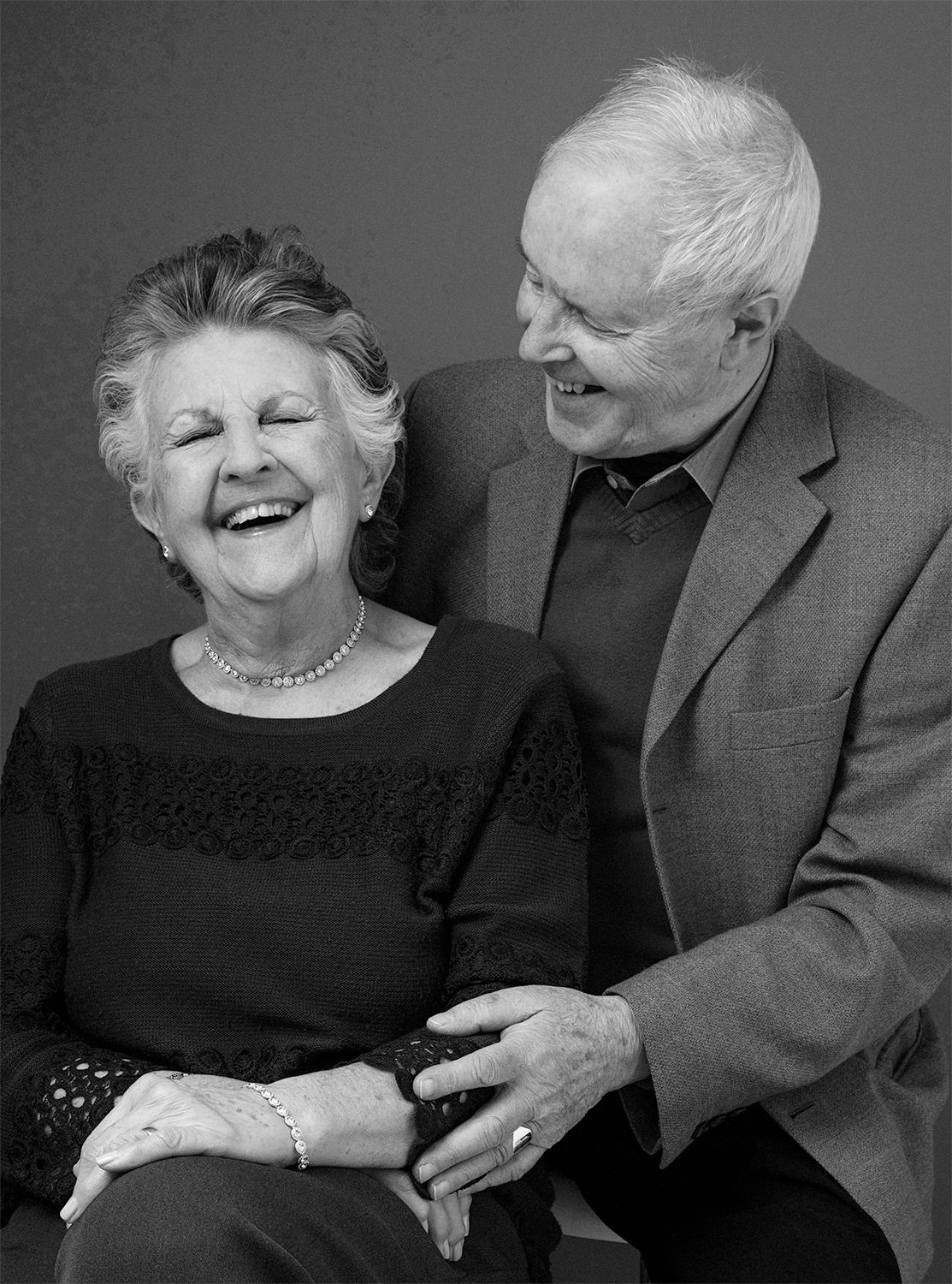 Black and white portrait of elderly couple laughing together against dark background.