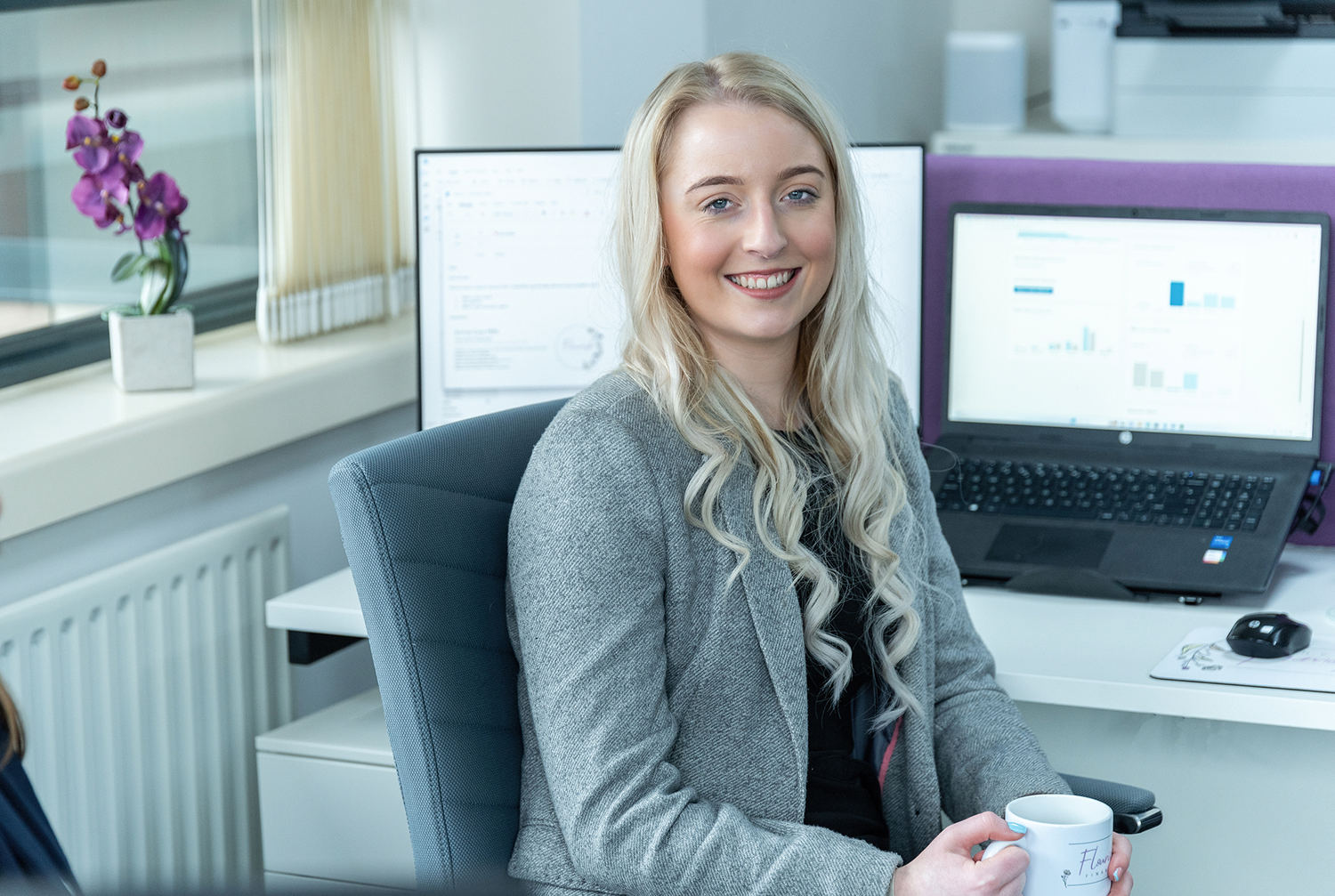 Smiling professional at desk with dual monitor computer setup and orchid decoration.