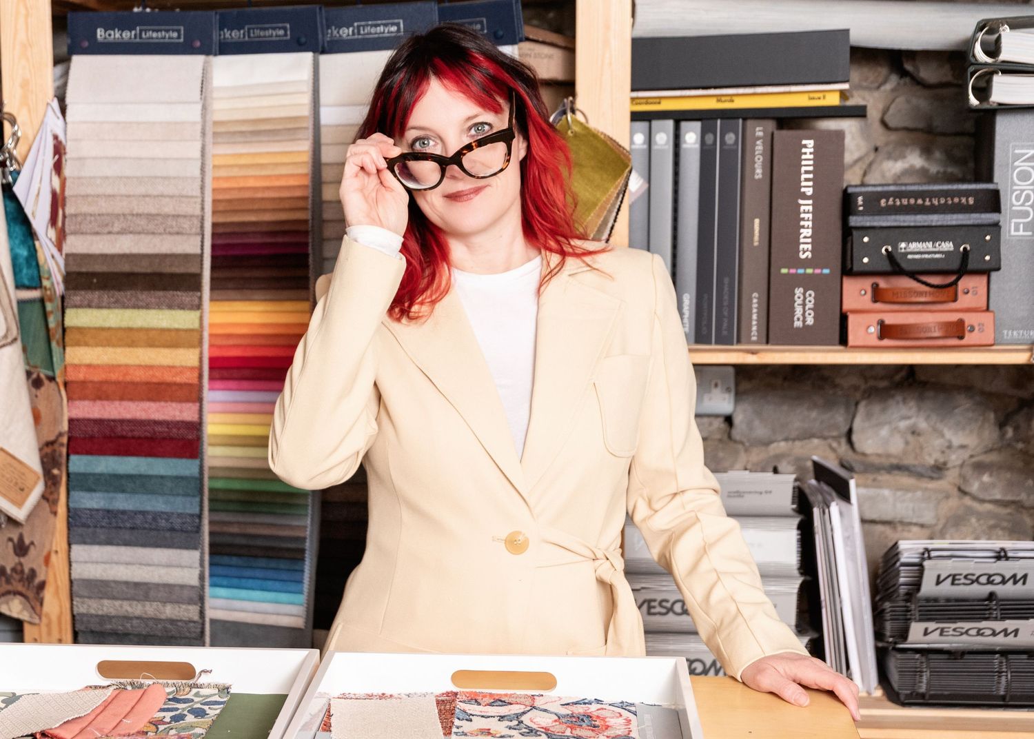 A person with red hair and glasses adjusts their frames while standing at a counter with fabric swatches and books.