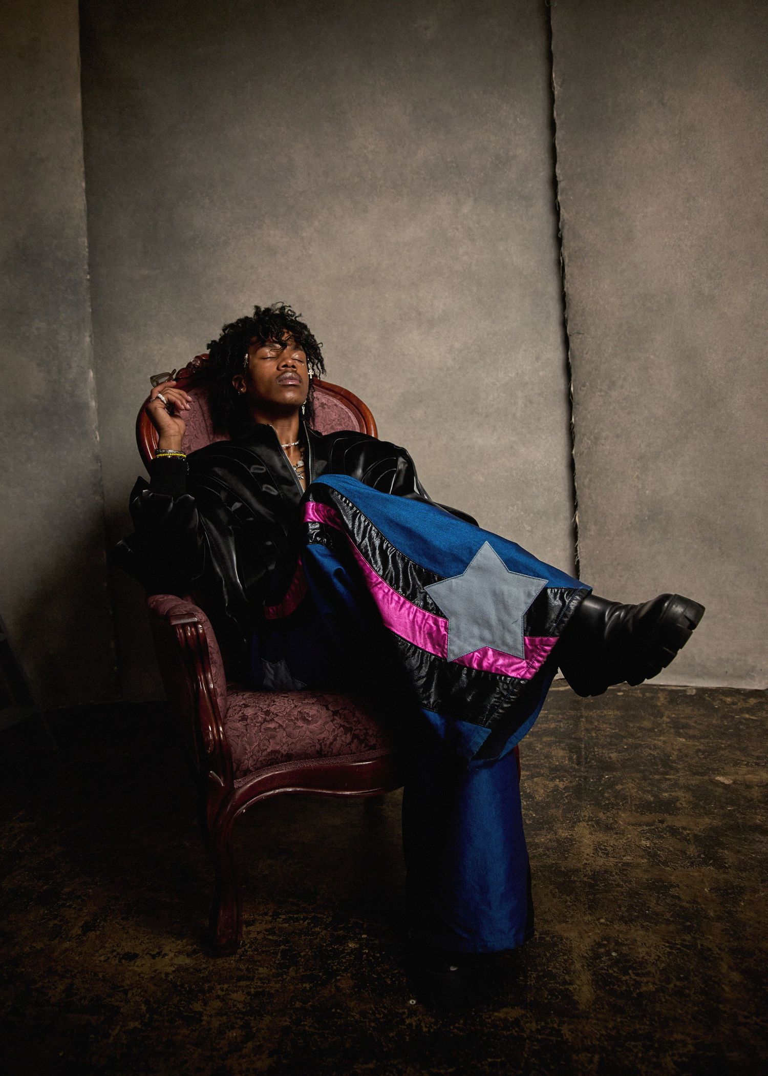 Stylish model lounging in leather armchair wearing black jacket and blue pants with pink accents.