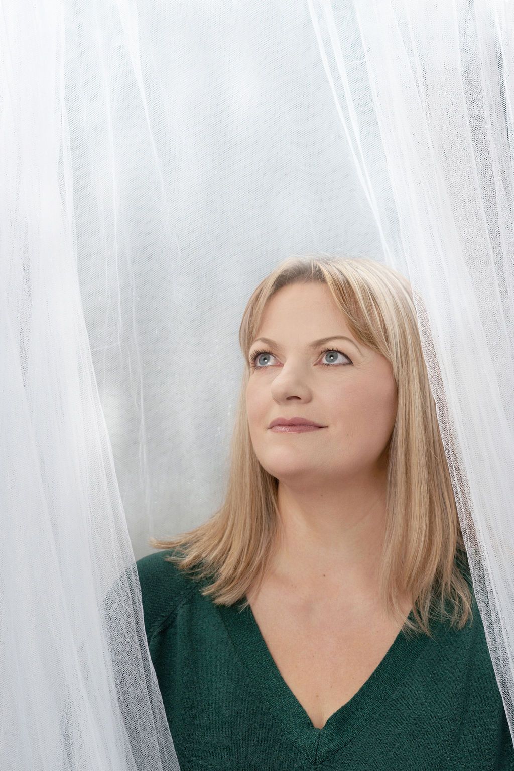Artistic portrait with blonde hair in dark green v-neck top against white flowing fabric background.