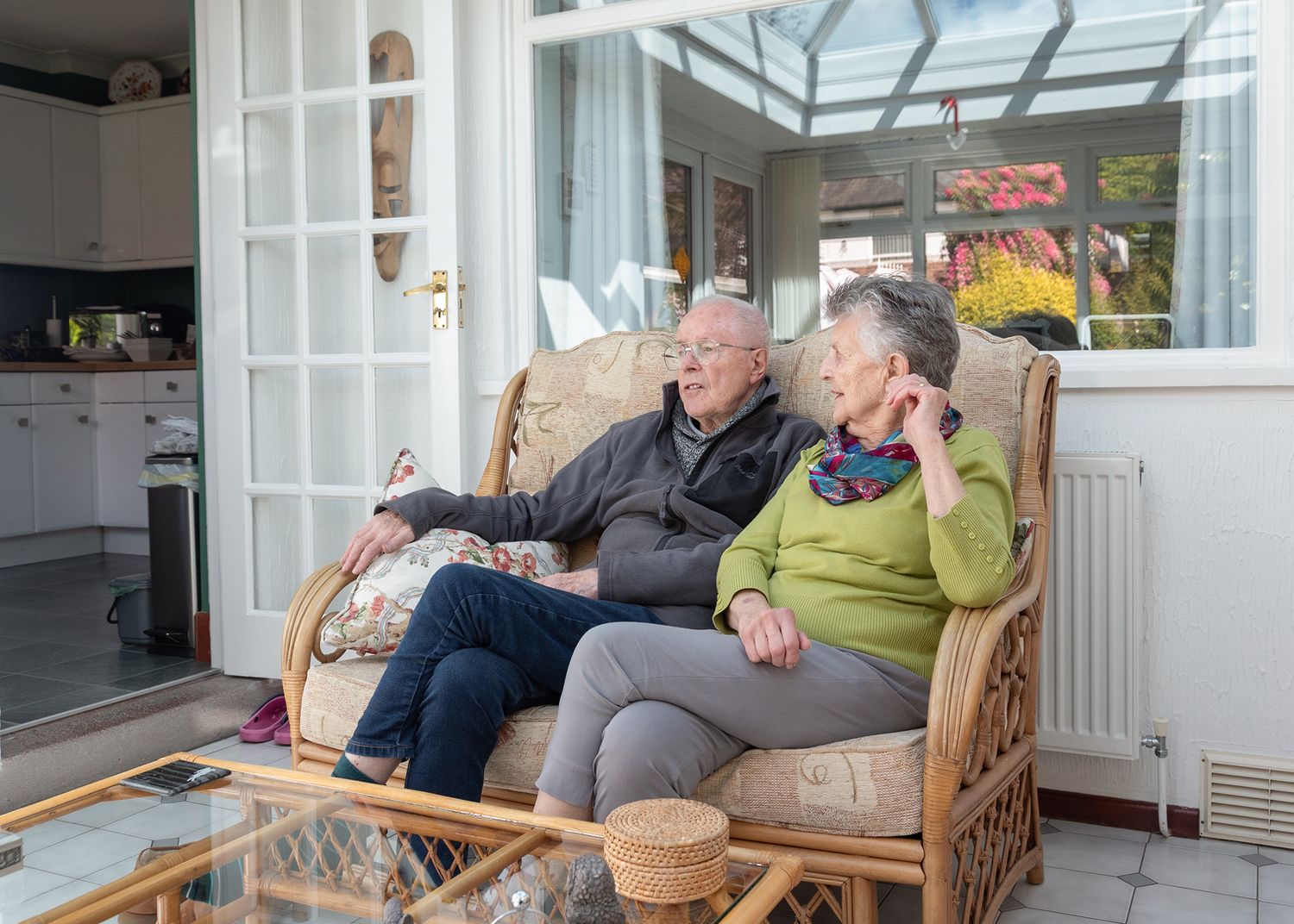 Elderly couple sitting together on a wicker sofa in a sunroom, relaxing and conversing at home.
